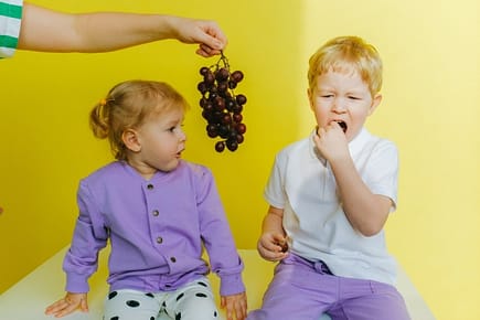 bambino e bambina mentre mangiando grappolo di uva tenuto da una mano adulta bambina e bambino esempio di invidia sfondo giallo mano con grappolo di uova al centro